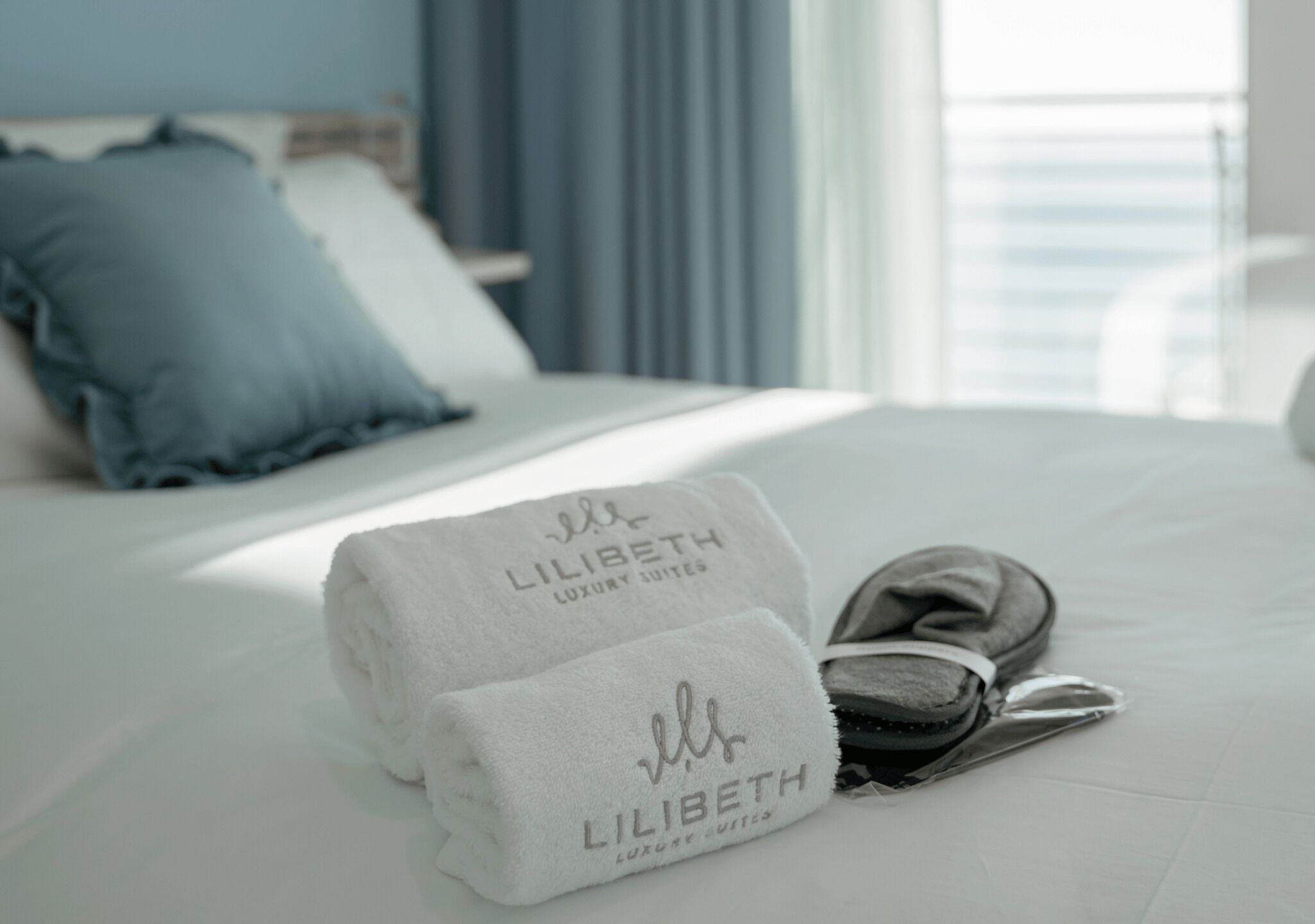 A neatly made bed with two towels and a pair of slippers, a blue pillow, and a window with curtains.