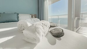 A room with a bed, two rolled towels, slippers, and a view of the sea through large windows.