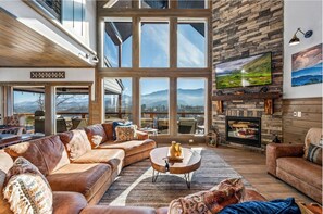 Un salon spacieux avec un grand canapé d’angle, une cheminée et une grande fenêtre donnant sur les montagnes.
