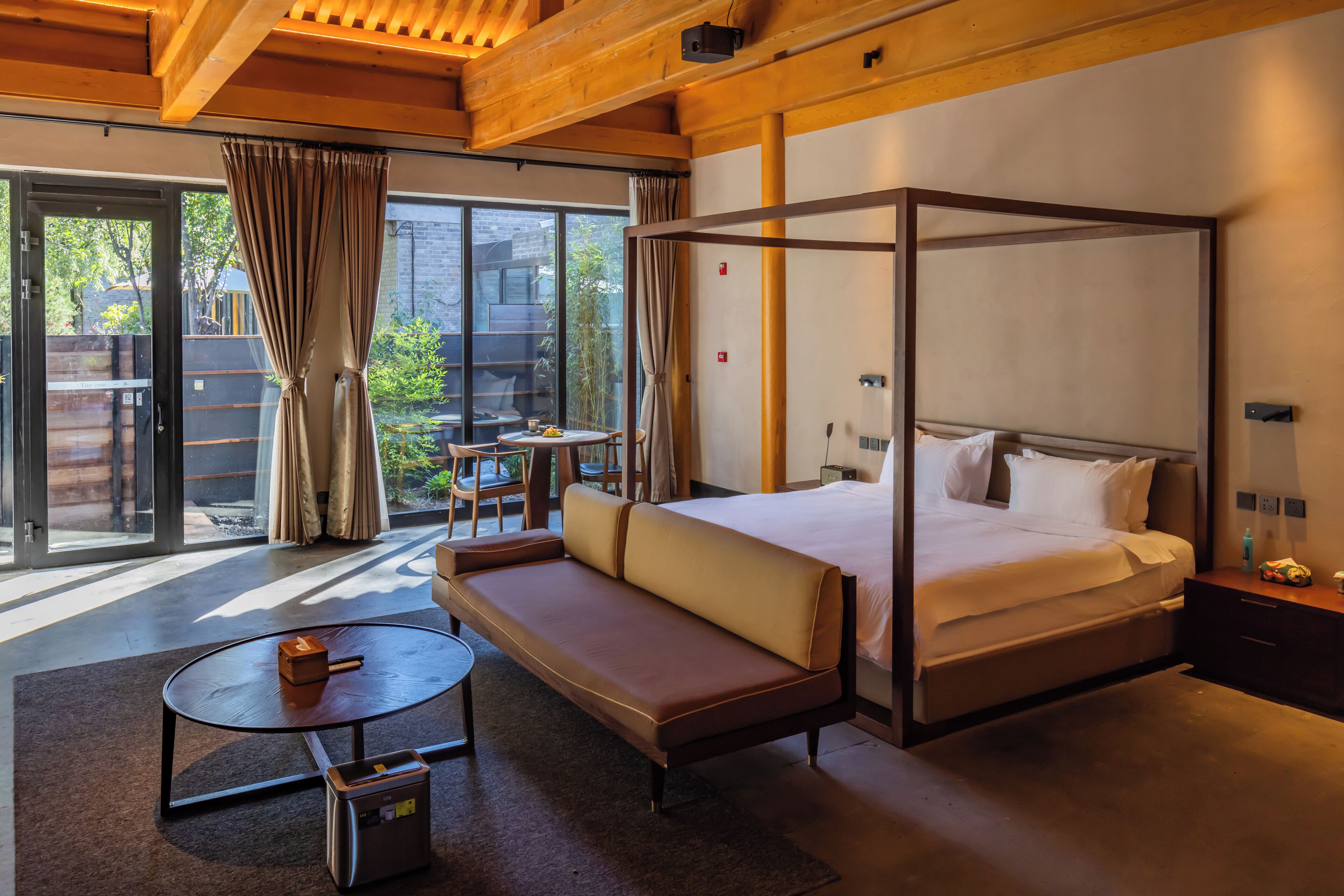 Habitación de hotel moderna con cama con dosel, un sofá, una pequeña mesa redonda y vistas al exterior a través de amplias ventanas.
