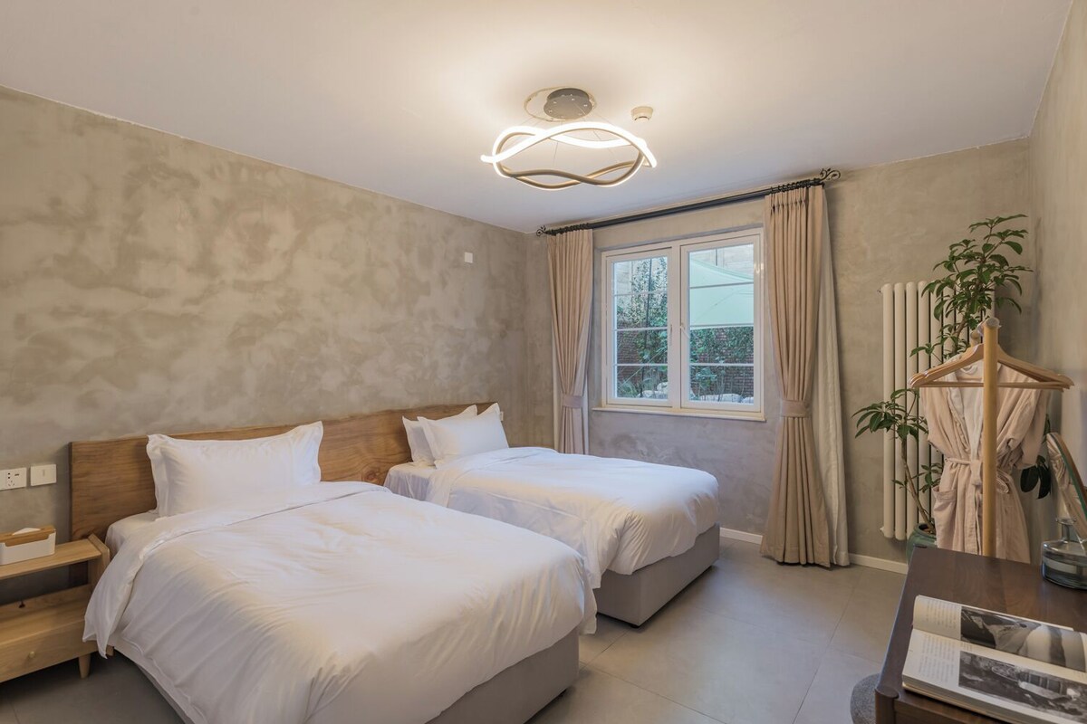 A modern hotel room with two beds, a wooden headboard, a large window with curtains, a radiator, and a plant.