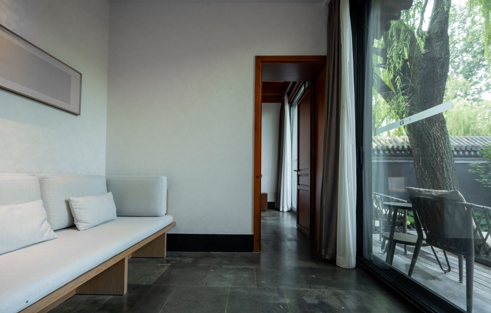 A modern interior with a sofa, a wooden bench, a large window with curtains, and a view of a tree and outdoor area.