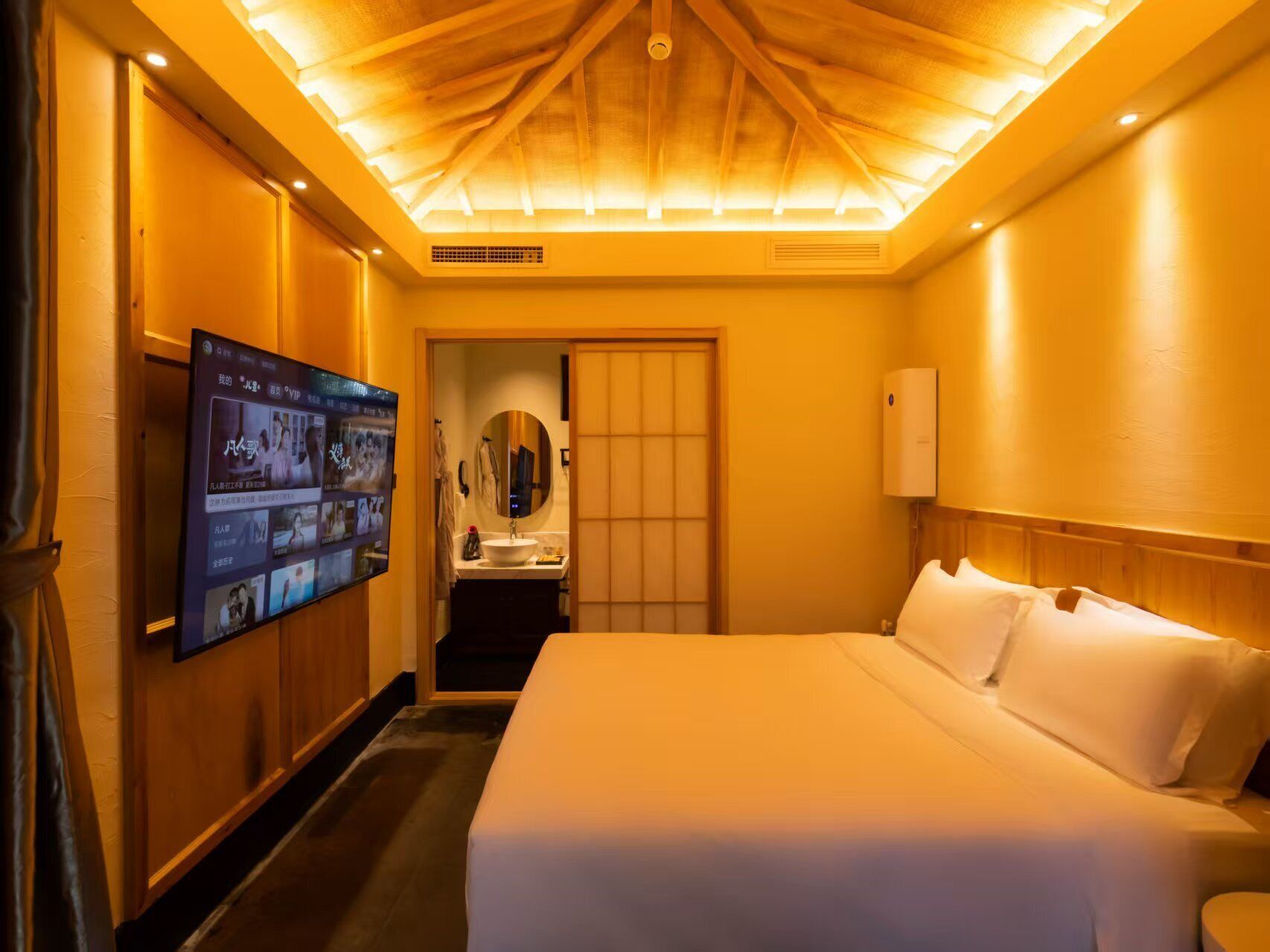 A hotel room with a large bed, a television mounted on the wall, a bathroom with a sink and mirror, and a ceiling with recessed lighting.