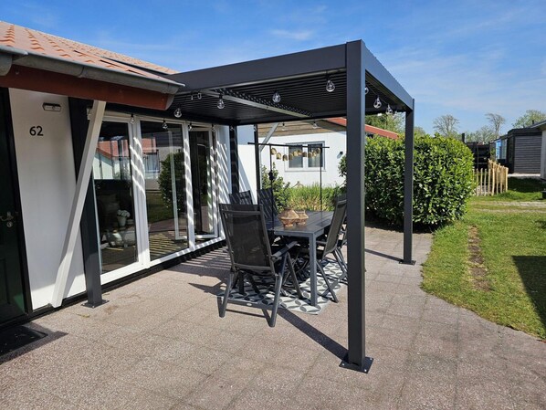 A covered patio area with a table and chairs, a pergola with hanging lights, and a house number 62. - Bungalow in Park With Fenced Garden (Sint Maartensvlotbrug)