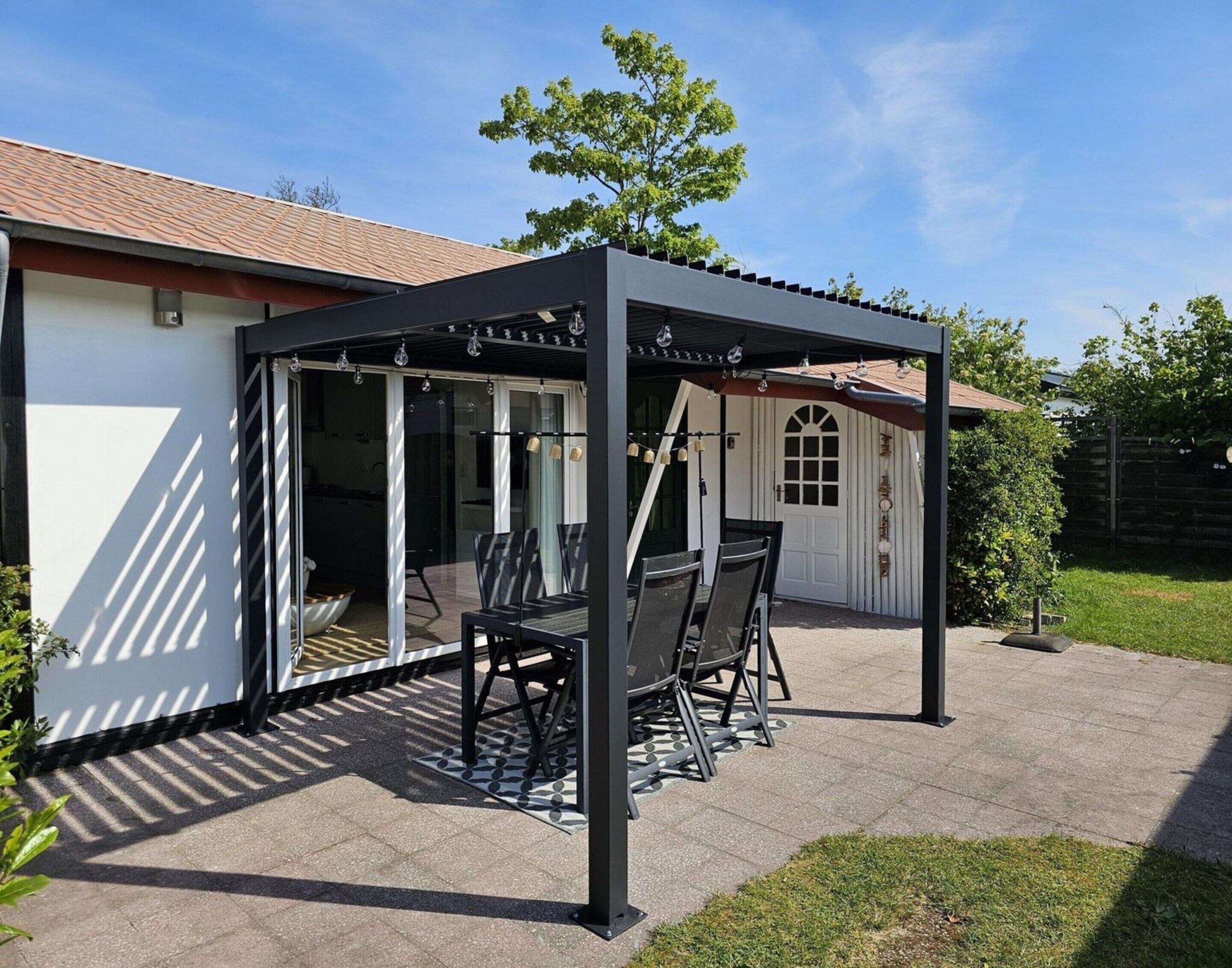 Katettu terassialue, jossa on pöytä ja tuoleja, pergola, jossa on valonauhat, ja taustalla valkoinen rakennus, jonka katto on päällystetty tiilillä.