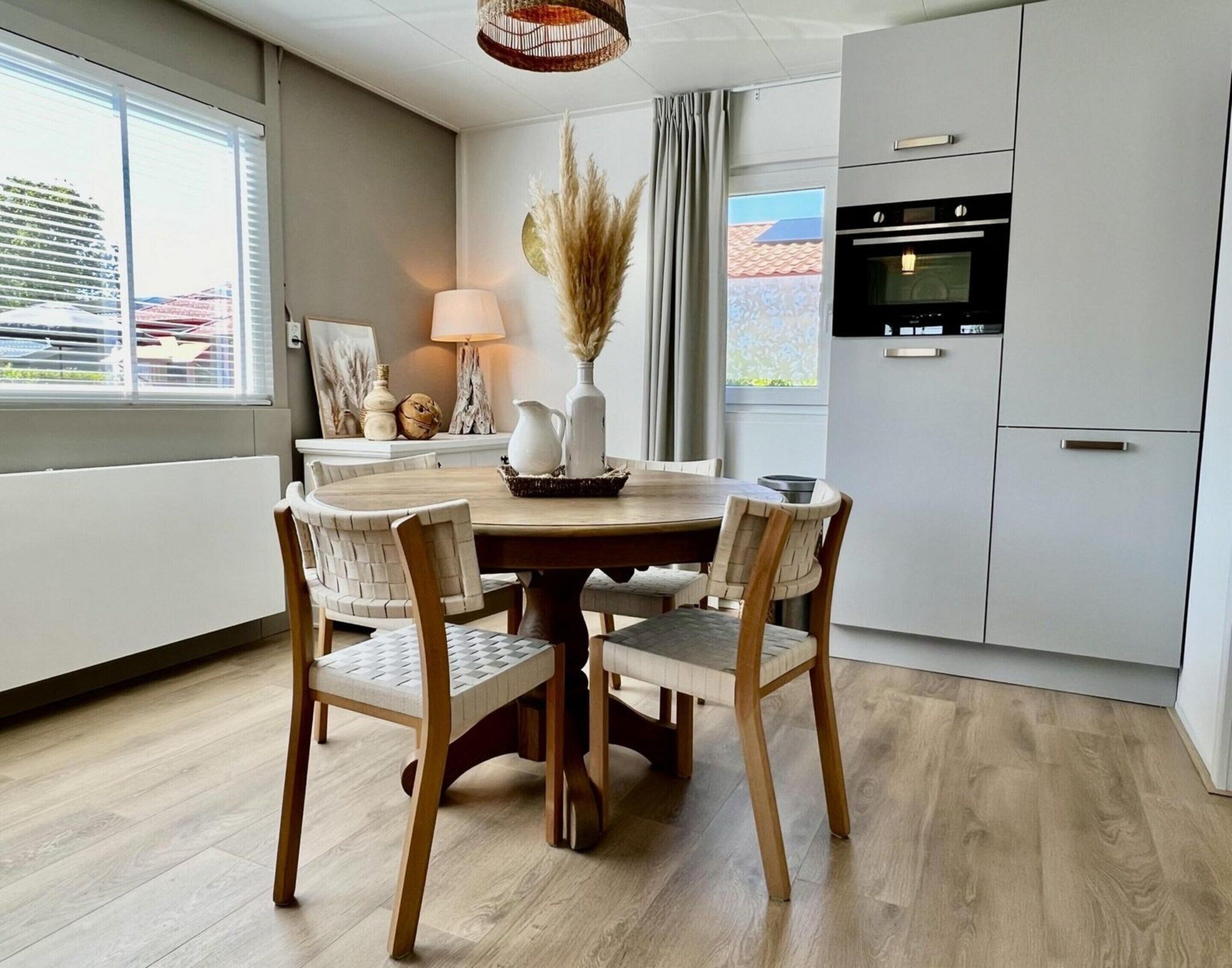 A modern kitchen with a round dining table, wooden chairs, a built-in oven, and a view of a residential area through the window.
