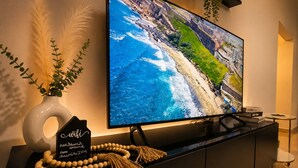 Ein modernes Wohnzimmer mit einem großen Flachbildfernseher an der Wand, einer schwarzen Unterhaltungseinheit und Dekorationselementen, darunter eine Vase mit Pflanzen und eine Lichterkette aus Holzperlen.