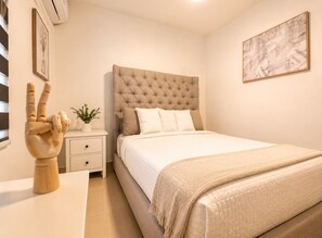Un dormitorio con cabecera capitonada, una mesita de noche con una planta, aire acondicionado de pared y un cuadro enmarcado.