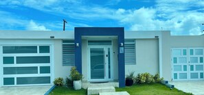 A modern two-story house with a garage, a front door, and a small front yard with potted plants. - Family Home • Free Parking • WIFI • Near Airport & Beaches (San Juan)