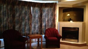 A living room with a fireplace, two armchairs, a small table, and a large window with curtains.