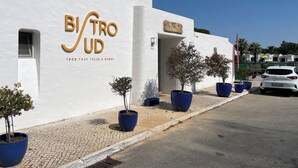 Un edificio con fachada blanca y un letrero que dice 'Bistro Ud' con el eslogan 'Comida que cuenta una historia'. Hay plantas en macetas y un coche blanco estacionado adelante.