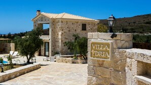 Une villa en pierre avec un toit en tuiles, une enseigne indiquant « Villa Ydria », et un espace piscine avec des chaises longues.