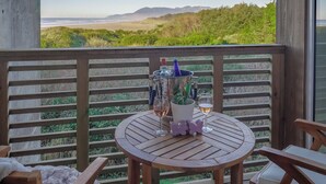 Una terraza de madera con una mesa redonda y sillas, con vistas a una playa y colinas.