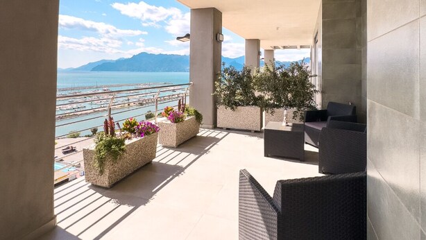 Un balcón amplio con vistas a un puerto deportivo y montañas, que cuenta con mobiliario exterior y macetas con flores.