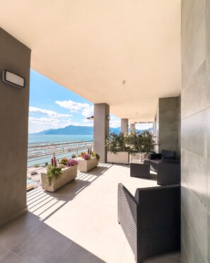 Un balcón amplio con vistas a un puerto deportivo y montañas, que cuenta con mobiliario exterior y macetas con flores.
