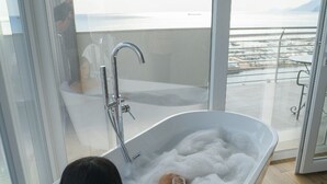 Una mujer se relaja en una bañera blanca llena de burbujas, sosteniendo una copa de vino blanco, con vistas al mar y las montañas a través de amplios ventanales.