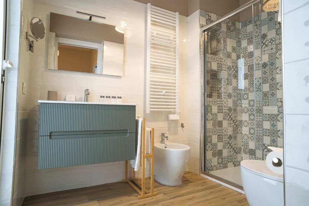 Un baño moderno con suelo de madera, un mueble de tocador azul, un bidé blanco y una ducha con azulejos estampados.
