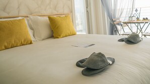 Un dormitorio con una cama grande, una lámpara de techo de ratán, vista al mar a través de una ventana y una mesa con sillas afuera.