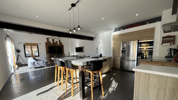 A modern kitchen with a central island, bar stools, a large refrigerator, and a dining area with a sofa and TV.