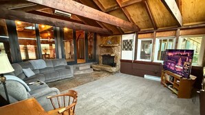 Una sala de estar acogedora con chimenea de piedra, un sofá gris, un sillón de madera y un televisor sobre un mueble de madera.