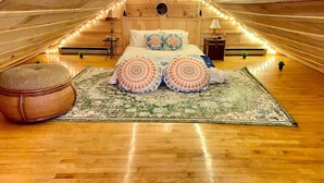 An attic bedroom with a wooden sloped ceiling, a bed with patterned pillows, a round ottoman, and a large rug. - Penthouse w/ Boho Loft, Jacuzzi, + Balcony -3 BR (Saint Joseph)
