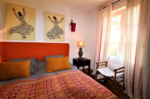 A bedroom with a red headboard, a patterned bedspread, a nightstand with a lamp, and a chair by the window.