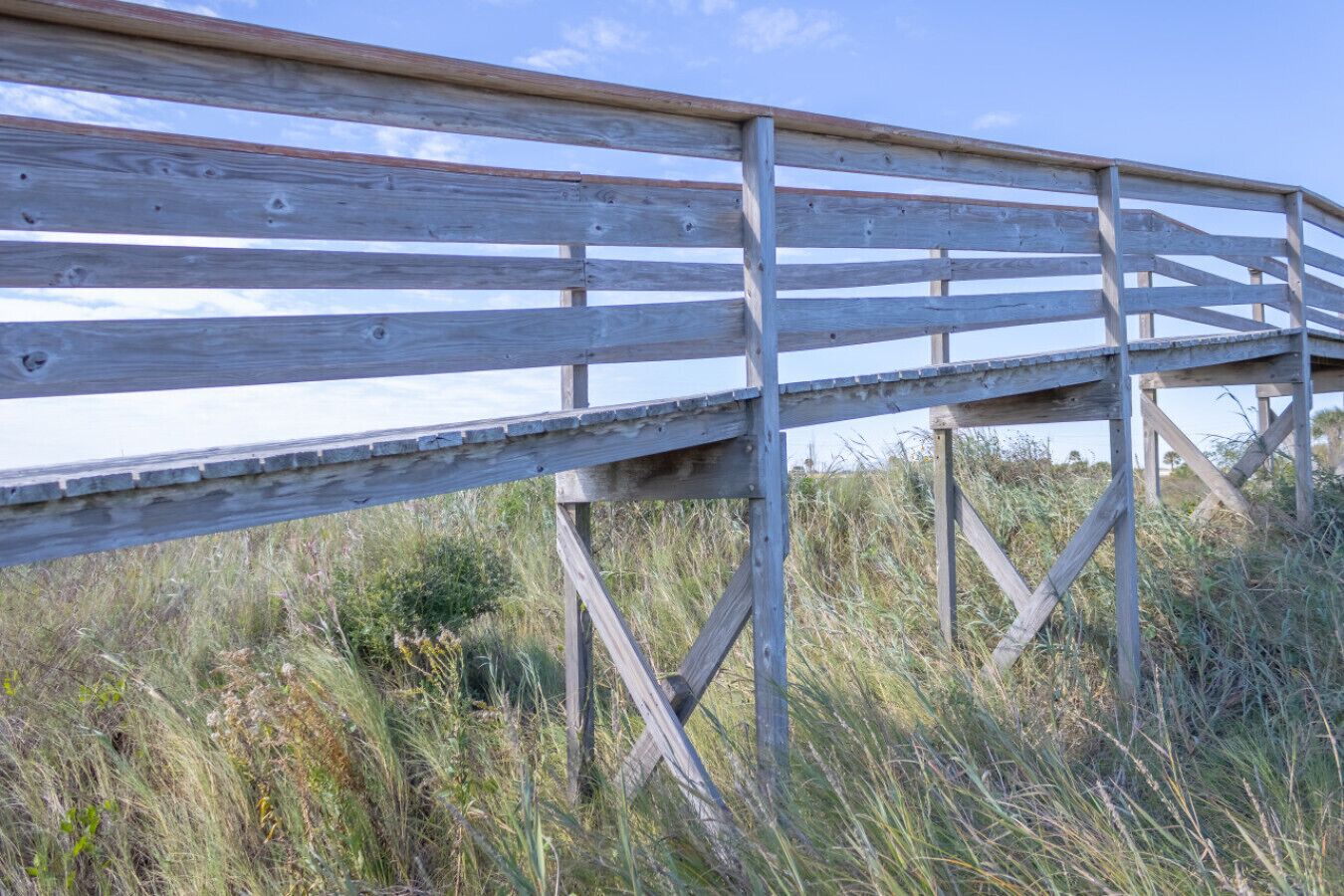Een houten looppad met een metalen draagconstructie, omringd door hoog gras en begroeiing.