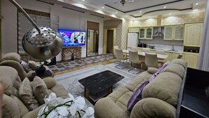 A modern living room with a sofa, coffee table, and a TV. There is a kitchen area with cabinets and a dining table. - ZimaLeto (Yusufhona)