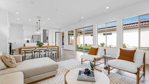 Una sala de estar moderna con sofá, mesa de centro y sillas. Hay un comedor con mesa y sillas. Grandes ventanales ofrecen vistas al exterior.
