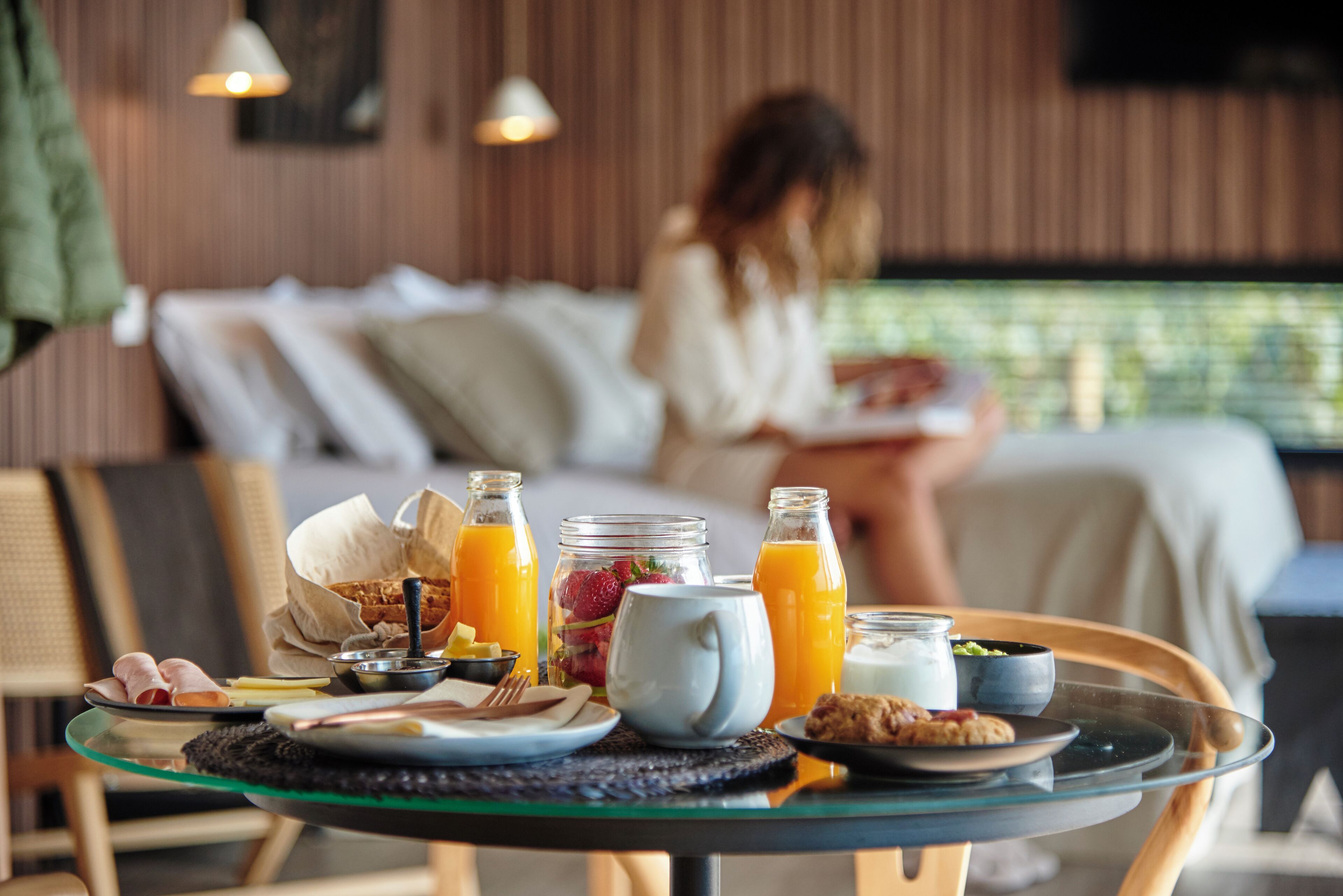 A breakfast setup with a glass table, plates of food, and a woman in the background.