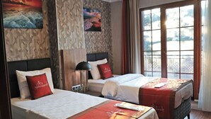 A hotel room with two beds, each with a red and white bedspread, a headboard, and a nightstand with a lamp. The room has wooden flooring, a large window with curtains, and a wall with a textured pattern and two paintings. - Marrakesch Hotel Tuzla (Tuzla)