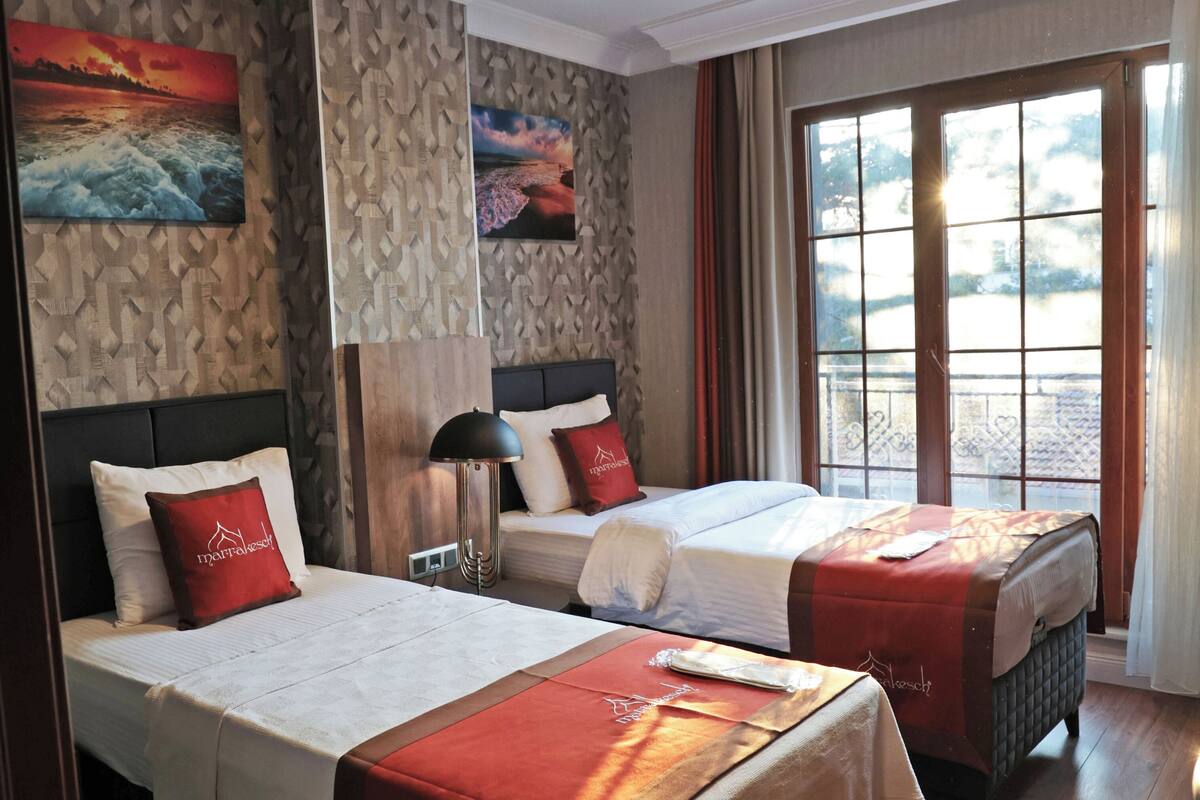 Une chambre d’hôtel avec deux lits, chacun recouvert d’une couvre-lit rouge et blanche, doté d’une tête de lit et agrémenté d  ’une table de chevet avec une lampe. La chambre est pourvue d’un plancher en bois, d’une grande fenêtre avec des rideaux et d’un mur orné d’un motif texturé et de deux tableaux.