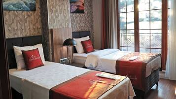 Ein Hotelzimmer mit zwei Betten, jeweils mit einer roten und weißen Bettdecke, einem Kopfteil und einem Nachttisch mit Lampe. Das Zimmer verfügt über Holzböden, ein großes Fenster mit Vorhängen sowie eine Wand mit strukturierter Oberfläche und zwei Bildern.