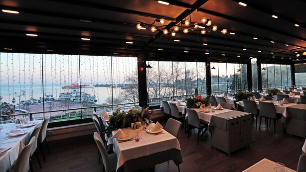 A restaurant with tables set for dining, large windows with a view of a body of water, and hanging lights on the ceiling. - Marrakesch Hotel Tuzla (Tuzla)