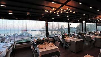 Ein Restaurant mit gedeckten Tischen, großen Fenstern mit Blick auf ein Gewässer und hängenden Leuchten an der Decke.