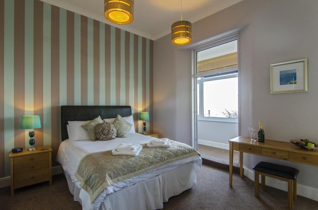 A hotel room with a large bed, two bedside lamps, a wooden desk with a bottle and glasses, and a window with a view.