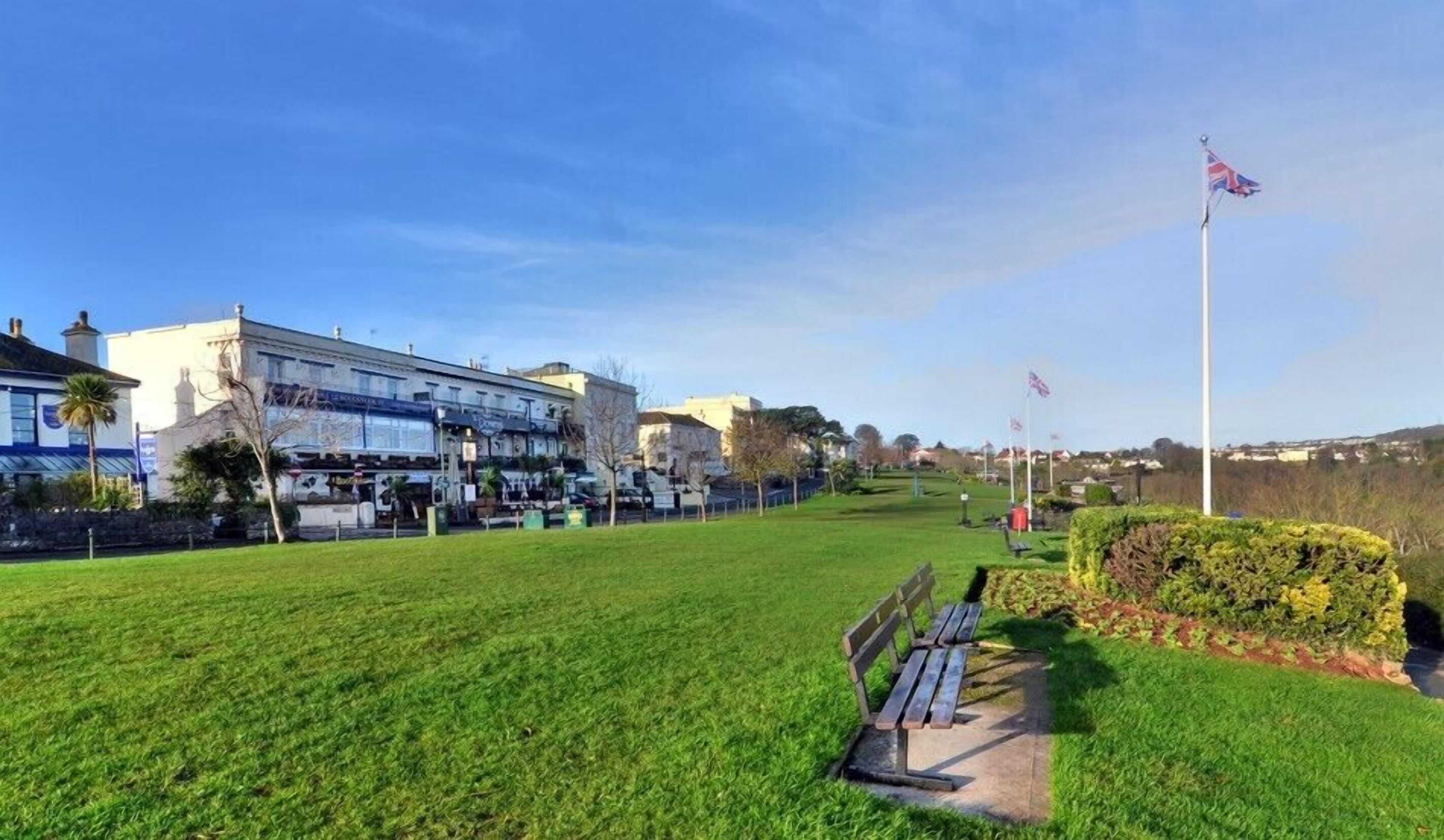 A grassy park area with a bench, a flagpole with flags, and a building with multiple stories and a blue sign.