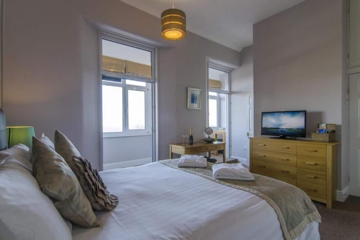 Ein Schlafzimmer mit einem großen Bett, einem Fernseher, einem Schminktisch und einem Fenster.