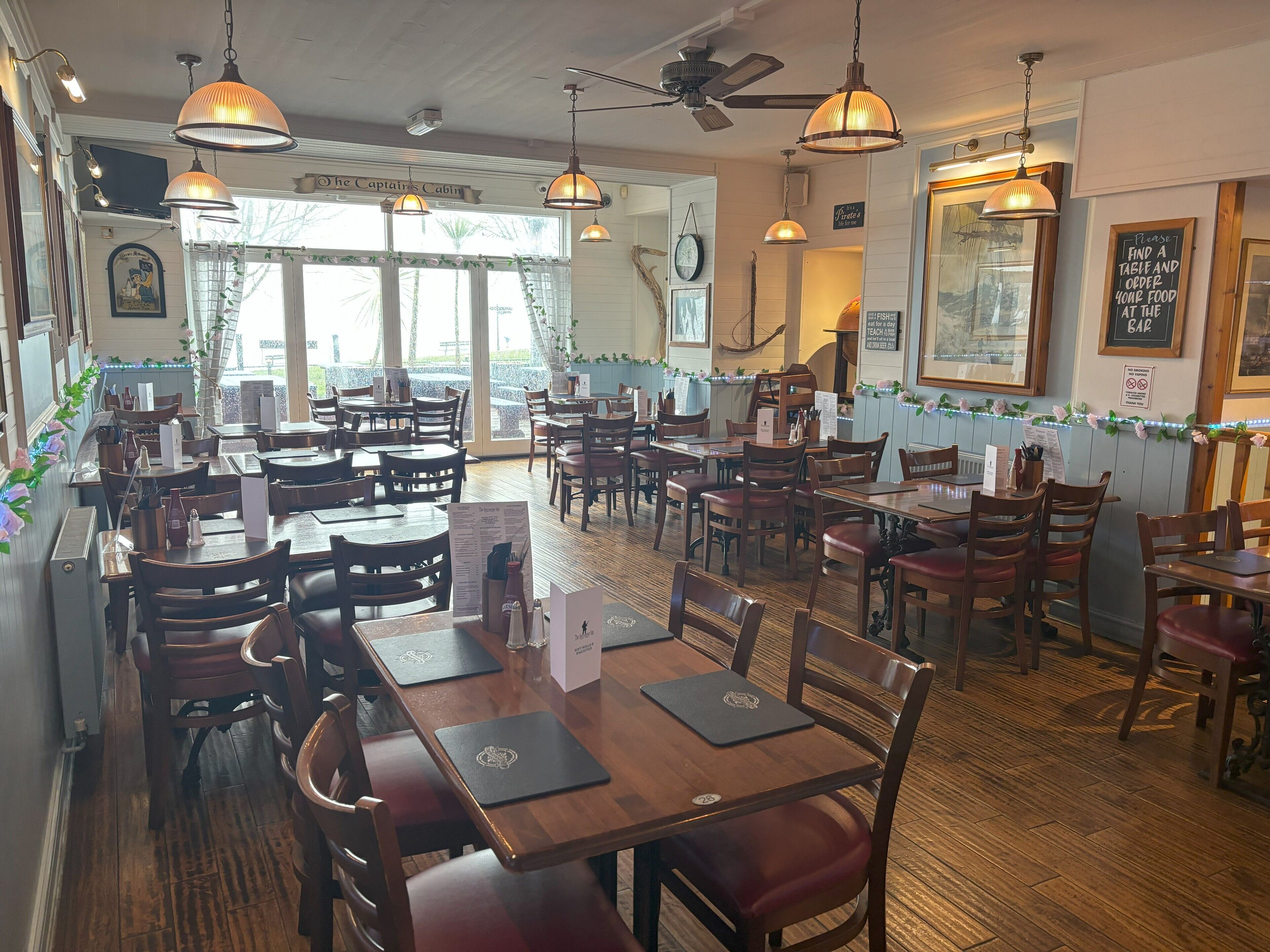 L’intérieur d’un restaurant avec des tables et des chaises en bois, des luminaires suspendus et une vue sur l’extérieur grâce à de grandes fenêtres.