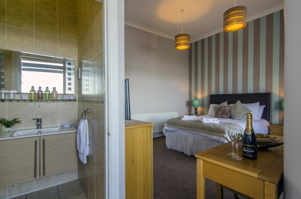 A hotel room with a bathroom, a bed with pillows, a bedside table with a bottle and glasses, and a striped wall.