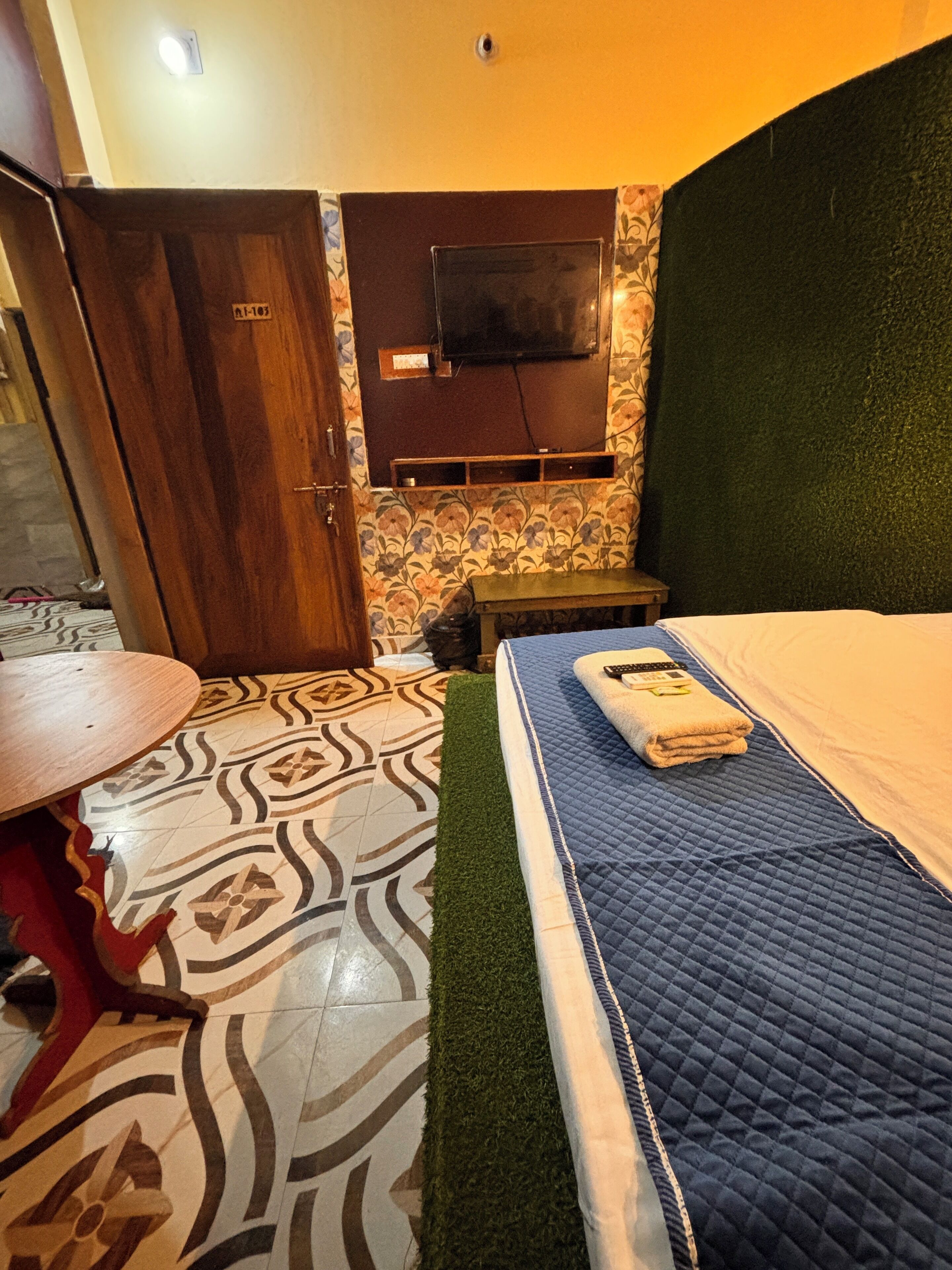 Una habitación con cama, una mesita pequeña, un televisor montado en la pared y piso estampado.