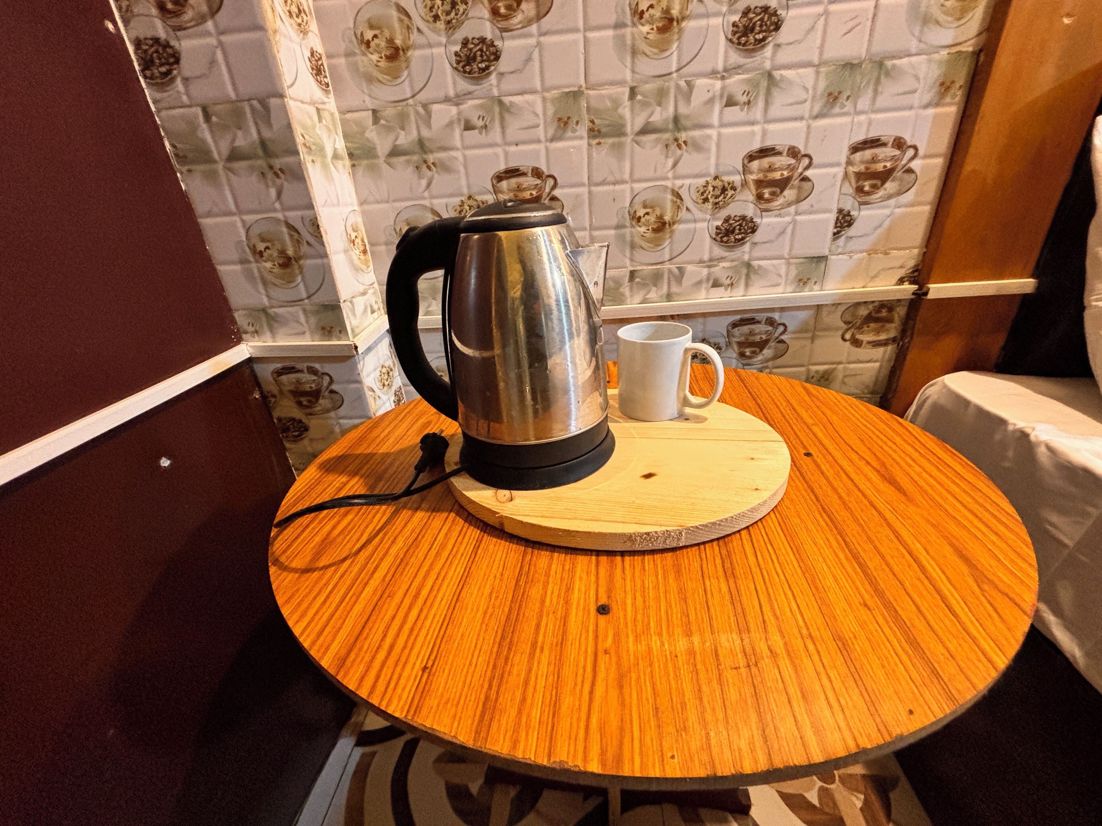 A round wooden table with a stainless steel kettle and a white mug on a coaster.