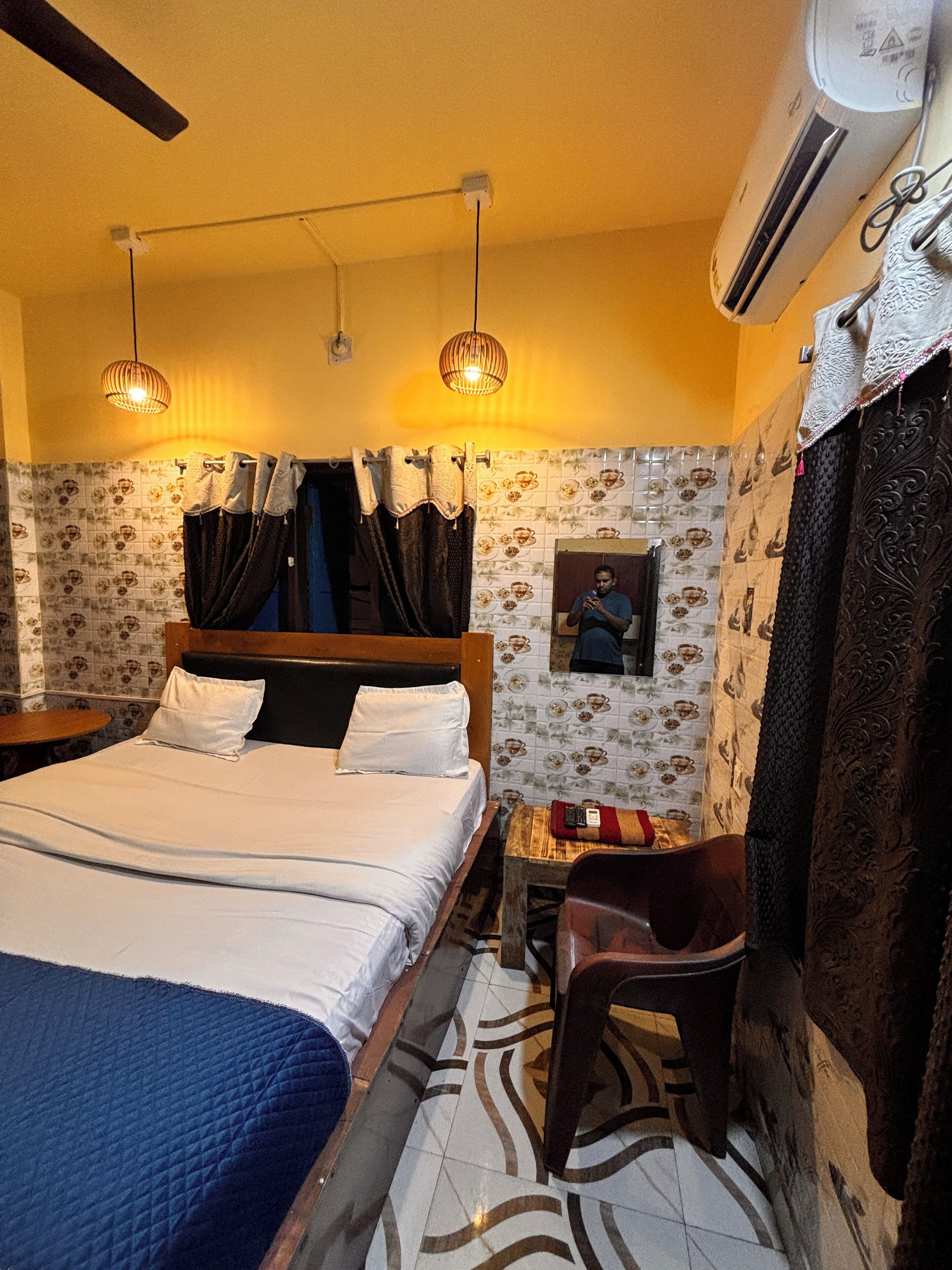 A hotel room with a bed, a small table, a chair, a mirror, and a wall-mounted air conditioner.