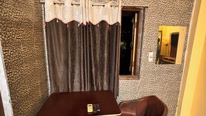 Ein kleiner Raum mit einem gemusterten Boden, einem Tisch mit Fernbedienung, einem Sessel, einer Klimaanlage und einem Fenster mit Vorhängen.