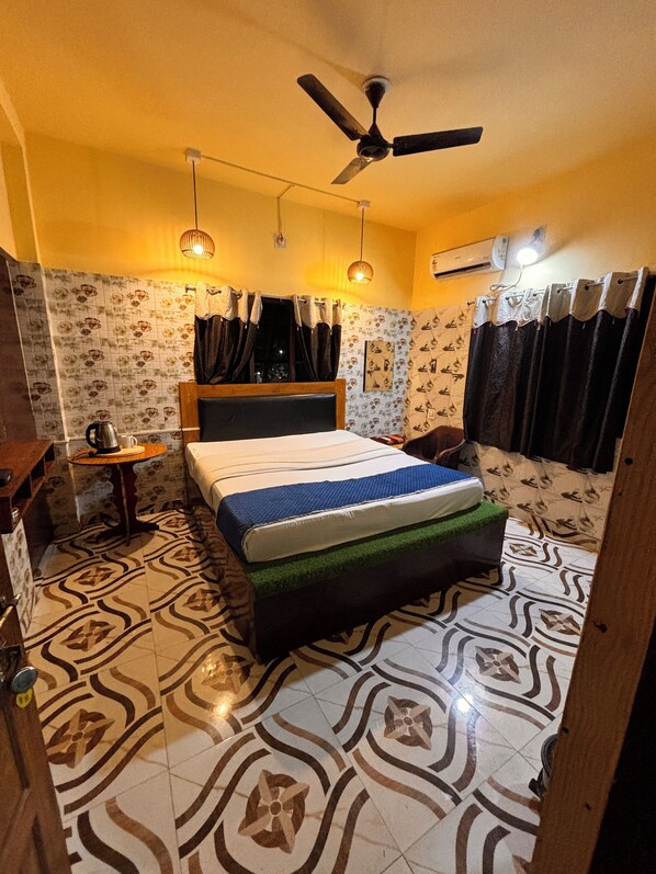 A bedroom with a bed, a ceiling fan, a small table with a kettle, patterned wallpaper, and a tiled floor.