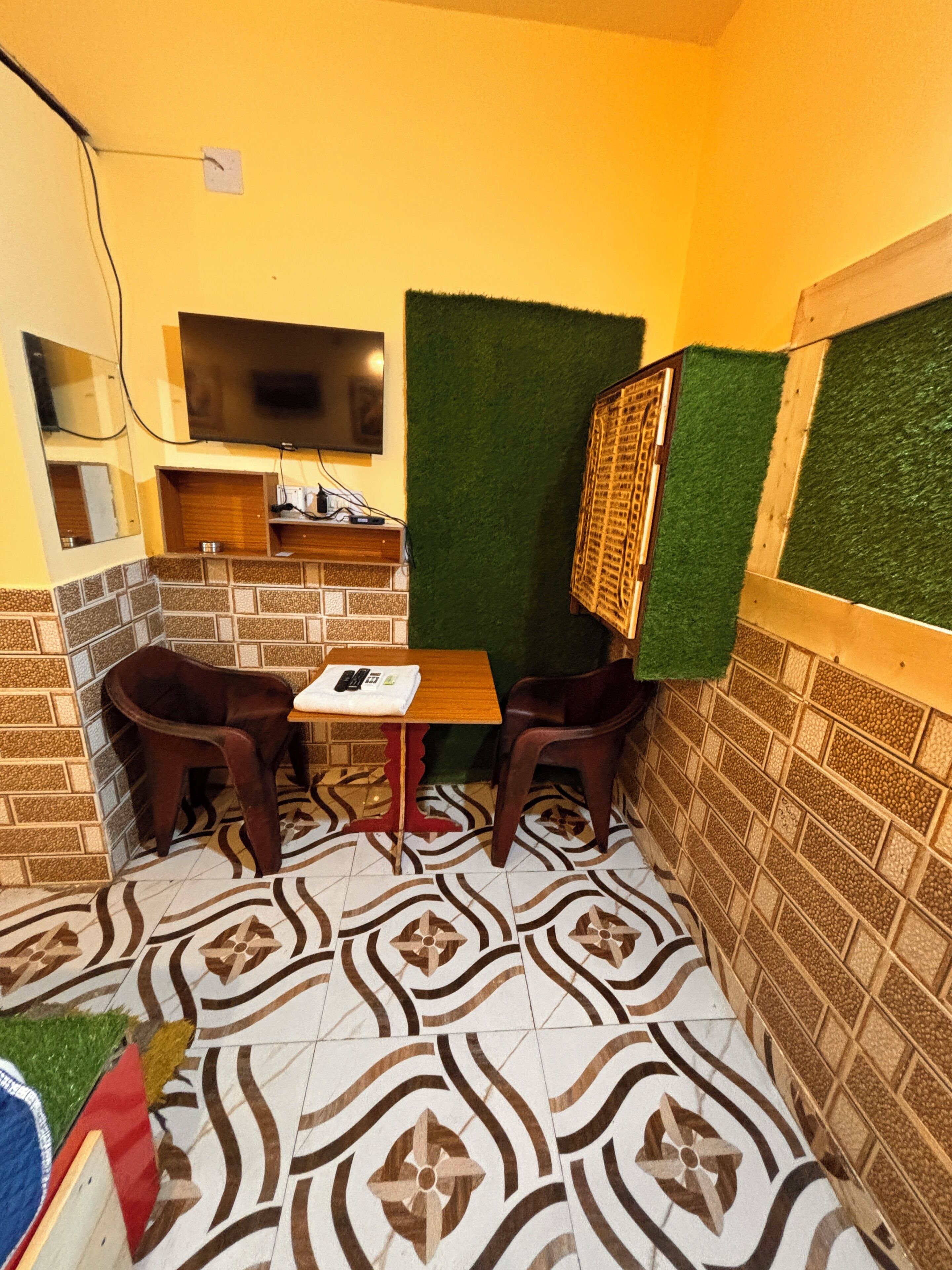 Una habitación pequeña y acogedora con piso de baldosas, un televisor montado en la pared, una pequeña mesa con dos sillas y un separador de césped artificial verde.