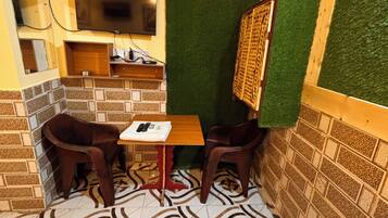 Una habitación pequeña y acogedora con piso de baldosas, un televisor montado en la pared, una pequeña mesa con dos sillas y un separador de césped artificial verde.