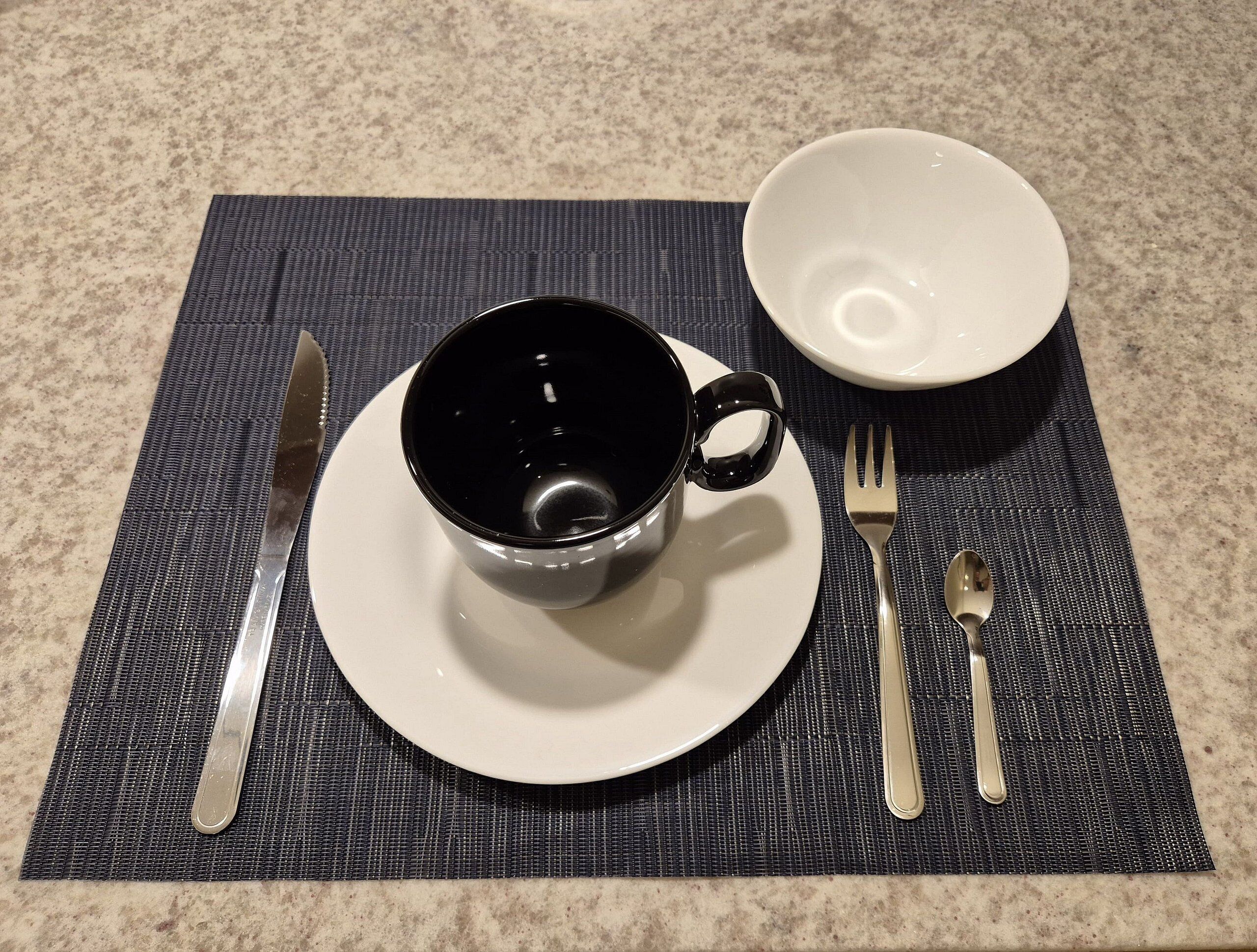 Une table dressée avec une tasse noire sur une assiette blanche, une fourchette et une cuillère posées sur un set de table foncé.