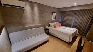 Een moderne slaapkamer met een grijze bank, een bed met wit en perzikkleurig beddengoed, een nachtkastje en een groot raam met gordijnen.