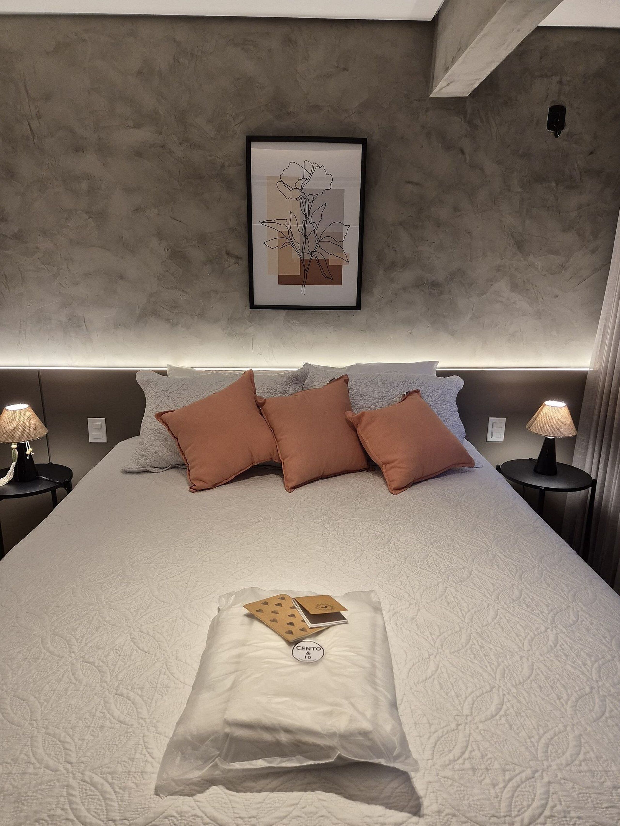 Cama bem arrumada com um edredom branco, duas almofadas castanhas e um cartão sobre a cama. Há uma obra de arte emoldurada na parede acima da cama.